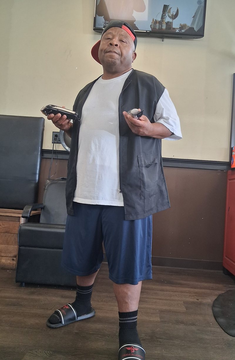 Tracy Tha Barber standing in his shop in a barber vest and cap, a pair of clippers in each hand.